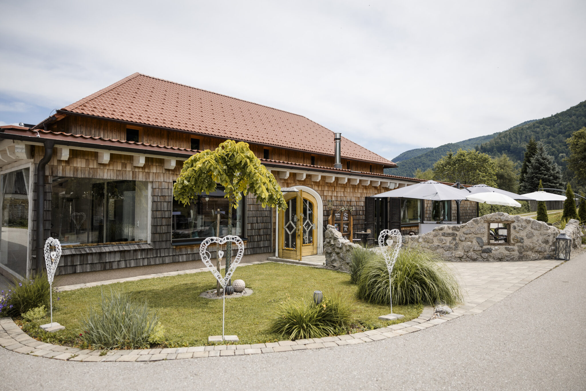 Eine bezaubernde Hochzeitslocation, Rieglergut, mit rotem Dach, herzförmigen Dekorationen und einer Terrasse mit Sonnenschirmen – perfekt für Ihre private Feier und mit Übernachtungsmöglichkeit.