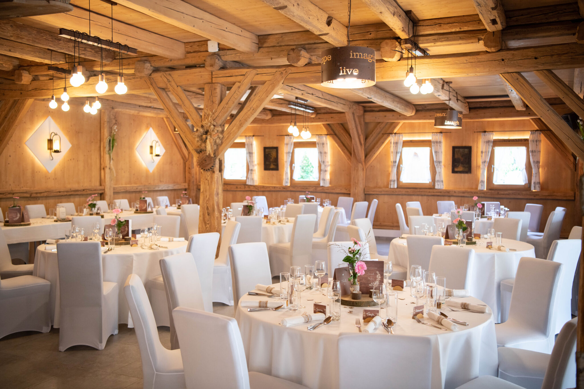 Ein rustikaler Festsaal mit Holzbalken, runden Tischen, weißen Stühlen und eleganter Tischdekoration – die perfekte Hochzeits- und Veranstaltungslocation mit bezauberndem Ambiente für jede Feier.