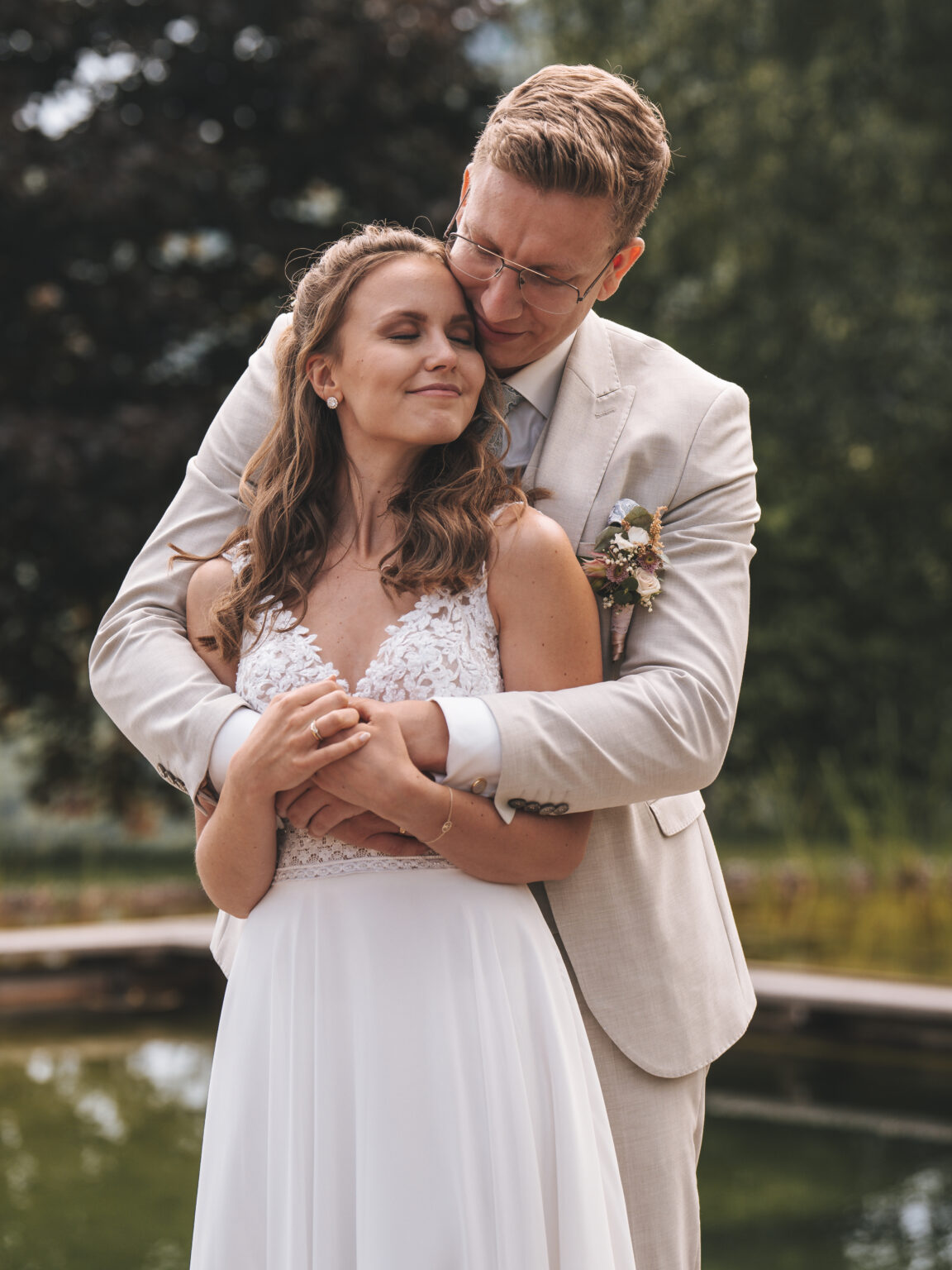 Ein Bräutigam in einem hellen Anzug umarmt seine lächelnde Braut in einem weißen Kleid im Freien in der Nähe eines Teiches an einer malerischen Hochzeitslocation.